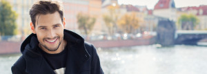 Young white man with black hair and neat beard and moustache, smiling by river's edge