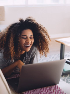 Laughing black woman, looking at her laptop while relaxing on her couch